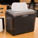 A black Just1 mini napkin dispenser on a table with white napkins next to a cup of coffee.