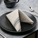 A white plate with a Hoffmaster FashnPoint black and white dishtowel print dinner napkin folded on it.