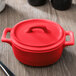 A red Bon Chef porcelain oval cocotte with a lid on a table.