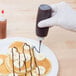A gloved hand uses a TableCraft squeeze bottle to drizzle chocolate syrup on a plate of pancakes with bananas and chocolate drizzle.