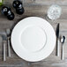A white Bon Chef bone china charger plate with silverware and a glass of water on a wooden table.