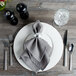 A Bon Chef white bone china charger plate with a napkin and silverware on a wooden table.