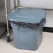 A Rubbermaid BRUTE grey square trash can with a lid on it next to a metal table.