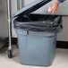 A person putting a black bag in a Rubbermaid BRUTE trash can with a lid.