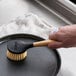 A person cleaning a pan with a Lodge scrub brush with a black and wooden handle.
