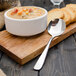A Walco 18/10 stainless steel bouillon spoon on a wood board next to a bowl of soup.