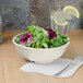 A bowl of salad with lettuce in a Thunder Group ivory melamine bowl on a table with a glass of water and a lemon wedge.