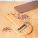 A wooden Bron Coucke chocolate shaver with a metal blade on a wooden surface with a chocolate bar and shavings.