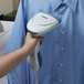 A person using a Conair white handheld steamer to steam a blue shirt.