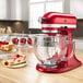 A KitchenAid candy apple red countertop mixer sits on a table with a slice of cheesecake on it.