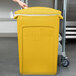 A person's hand holding a plastic bag over a yellow Rubbermaid Slim Jim trash can.