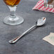 A Walco stainless steel iced tea spoon next to a glass of liquid on a table.
