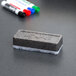 A grey rectangular Expo dry erase eraser with white foam on it next to several markers.