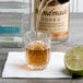 A person pouring a shot of brown liquid into an Arcoroc shot glass on a counter with a lime on a napkin.
