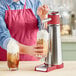A person wearing a red apron using a stainless steel iSi whipped cream dispenser to top a coffee drink.