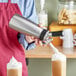 A person using an iSi stainless steel whipped cream dispenser to add whipped cream to a drink.