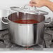 A hand using a Vollrath Optio sauce pot with a metal handle and lid on a stove.