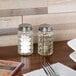 A Tablecraft glass jar with a stainless steel top containing salt and pepper on a table.