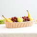 A Tablecraft woven rattan-like bread basket filled with bananas, apples, and fruit on a table.