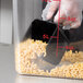 A hand using a Carlisle black polycarbonate portion scoop to measure macaroni into a container.