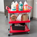 A red Rubbermaid three shelf utility cart with a variety of cleaning supplies on it.