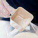 A gloved hand using a Carlisle beige polycarbonate portion scoop to fill a plastic container with rice.