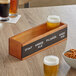An Acopa walnut finish flight crate with a drink and a bowl of pretzels on a table.