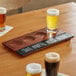 An Acopa walnut finish flight tray with four glasses of beer on it.