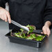A person using metal tongs to serve salad from a Cambro black polycarbonate food pan.