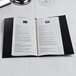 A black Menu Solutions wine list cover on a table with a white menu inside.