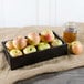 A Tablecraft black serving and display crate holding a box of apples and a glass jar of brown liquid.