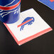 A blue and red Creative Converting Buffalo Bills beverage napkin with a buffalo logo on a table next to a cup with a buffalo logo.