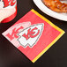 A Kansas City Chiefs beverage napkin with the team logo next to a plate of chicken.