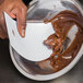 A person using an Ateco large plastic bowl scraper to mix chocolate frosting in a bowl.