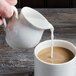 A hand pouring milk from a white Villeroy & Boch porcelain creamer into a cup of coffee.