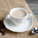 A white Villeroy & Boch porcelain cup filled with a foamy drink on a burlap surface with a spoon and coffee beans.