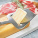 A Franmara stainless steel cheese plane slicing cheese on a metal surface.