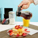 A person using a Tablecraft teardrop syrup dispenser to pour syrup on a stack of pancakes with strawberries.