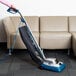 A person using a Lavex upright bagged vacuum cleaner with a blue and black handle to clean a couch.
