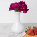 A white Arcoroc porcelain vase with purple flowers next to a plate of strawberries.
