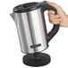 A close-up of a hand holding a silver and black Hamilton Beach electric kettle.