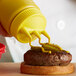 A person using a Vollrath Traex yellow bottle cap to put mustard on a burger.