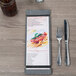 A customizable wood menu board with rubber band straps on a table with a menu and silverware.