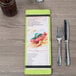 A Menu Solutions wood menu board with lime rubber band straps on a table with a menu, fork, and knife.