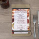 A Menu Solutions weathered walnut wood menu board on a table with a glass of brown liquid and silverware.