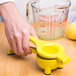 A hand using a Tablecraft yellow zinc alloy lemon squeezer to juice a lemon into a measuring cup.