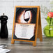 A Menu Solutions country oak wood sandwich menu board tent with picture corners on a table with a menu and wine glass.