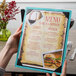A hand holding a Menu Solutions sky blue wood menu board with picture corners over a table in a fine dining restaurant.