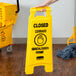 A hand holding a yellow Rubbermaid double sided wet floor caution sign.