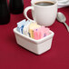 A white Tuxton China sugar packet holder on a counter with sugar packets in it.
